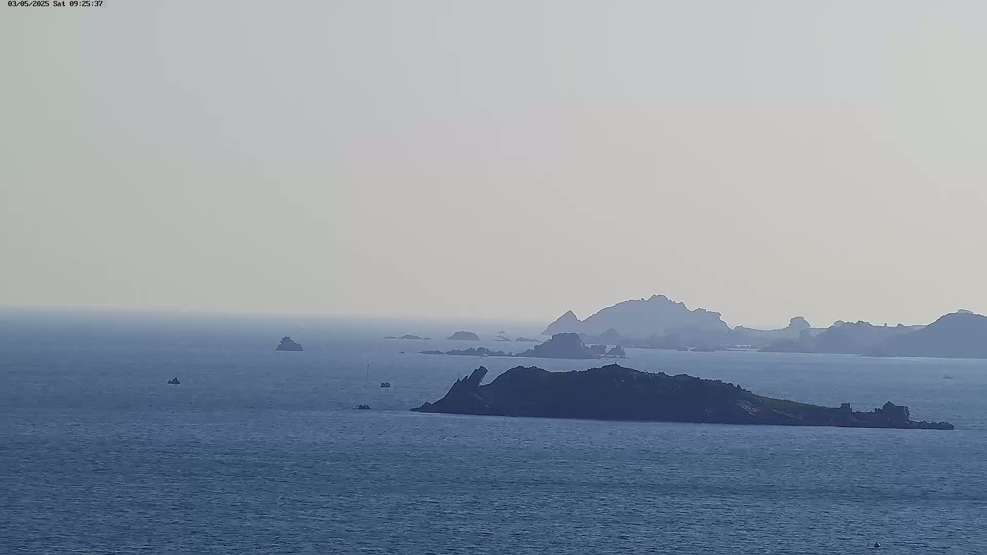 A hazy, distant view of several rocky islands and islets in a calm sea.