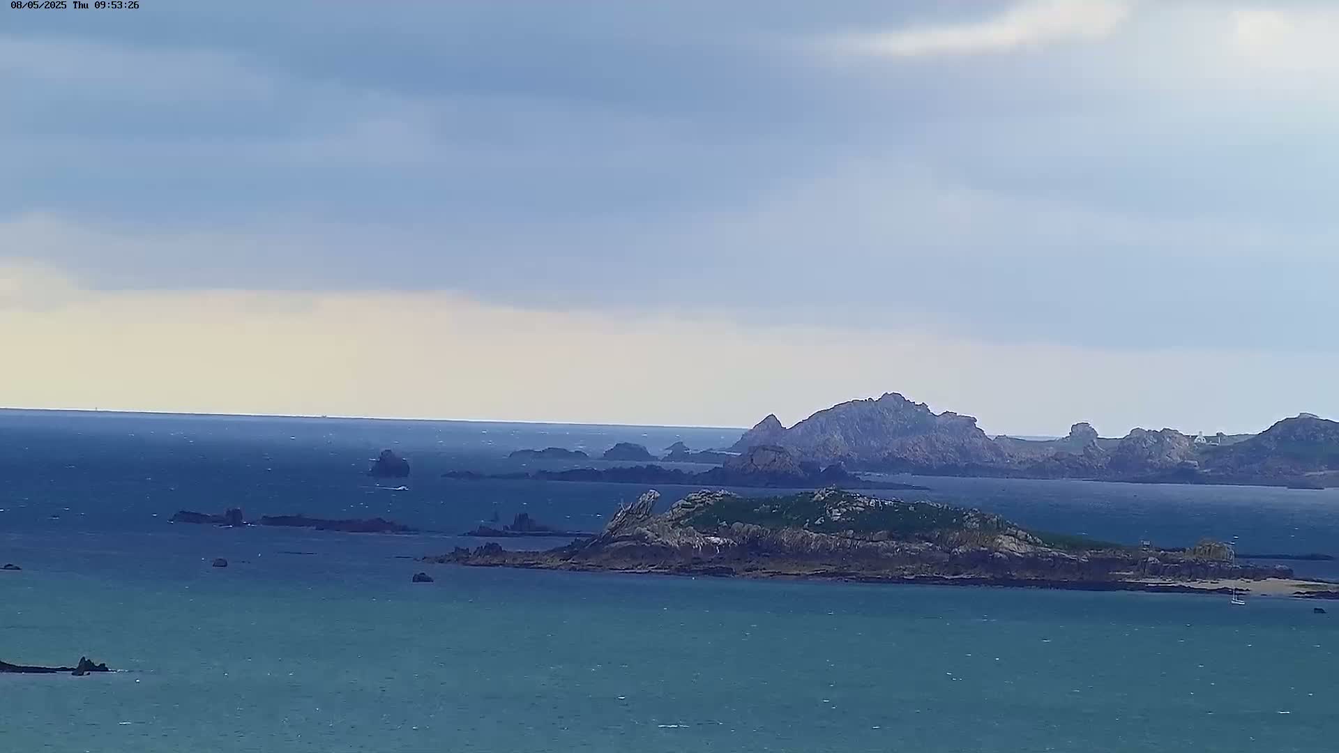 A tranquil seascape shows several rocky islands dotting a calm, blue-green ocean under a mostly cloudy sky.