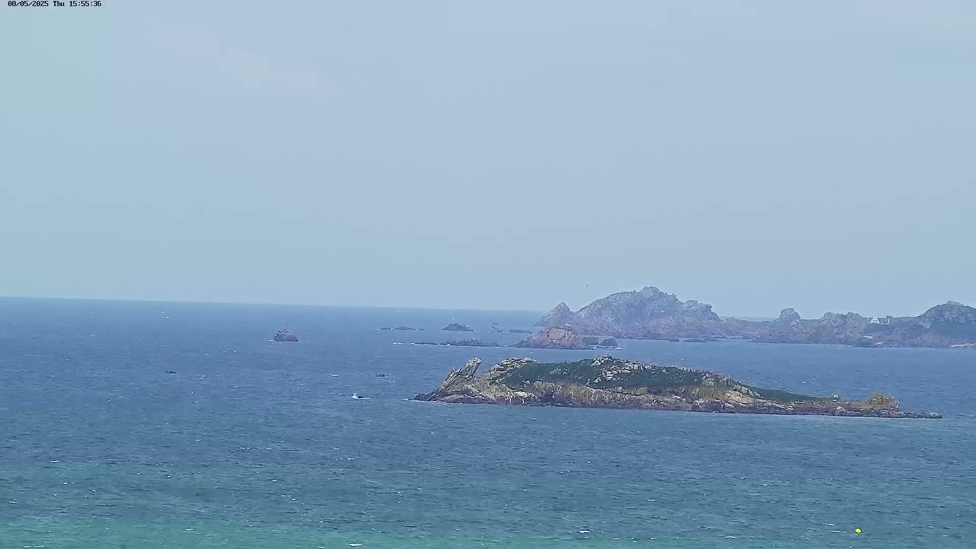 A hazy, overcast day reveals a rocky island in a vast, blue ocean with other smaller islands and rocks in the distance.