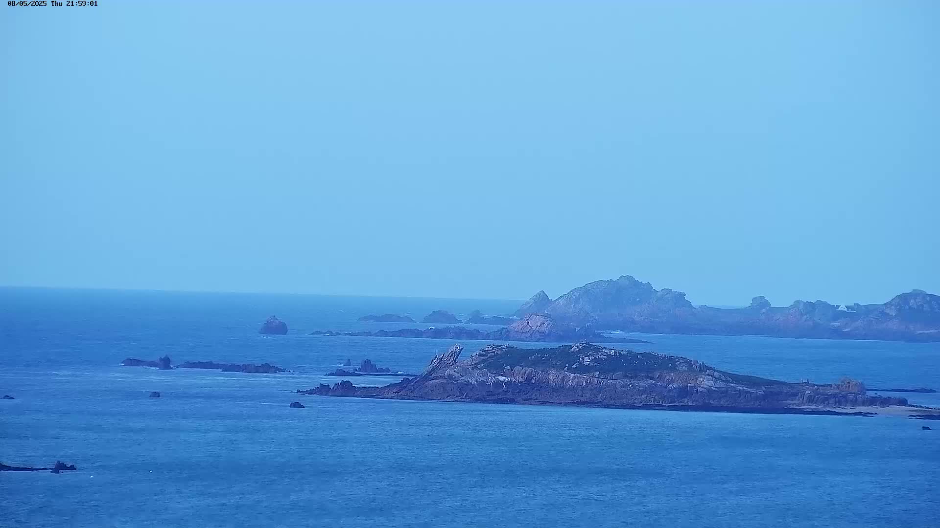 A tranquil, blue ocean dotted with numerous rocky islands under a clear, light-blue sky.