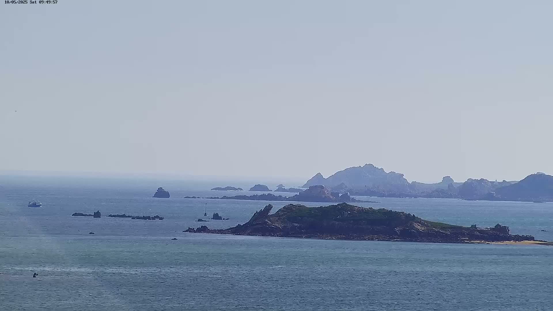 A hazy, sunny day reveals a calm ocean with several small rocky islands and a larger island with green vegetation, along with a few small boats in the distance.