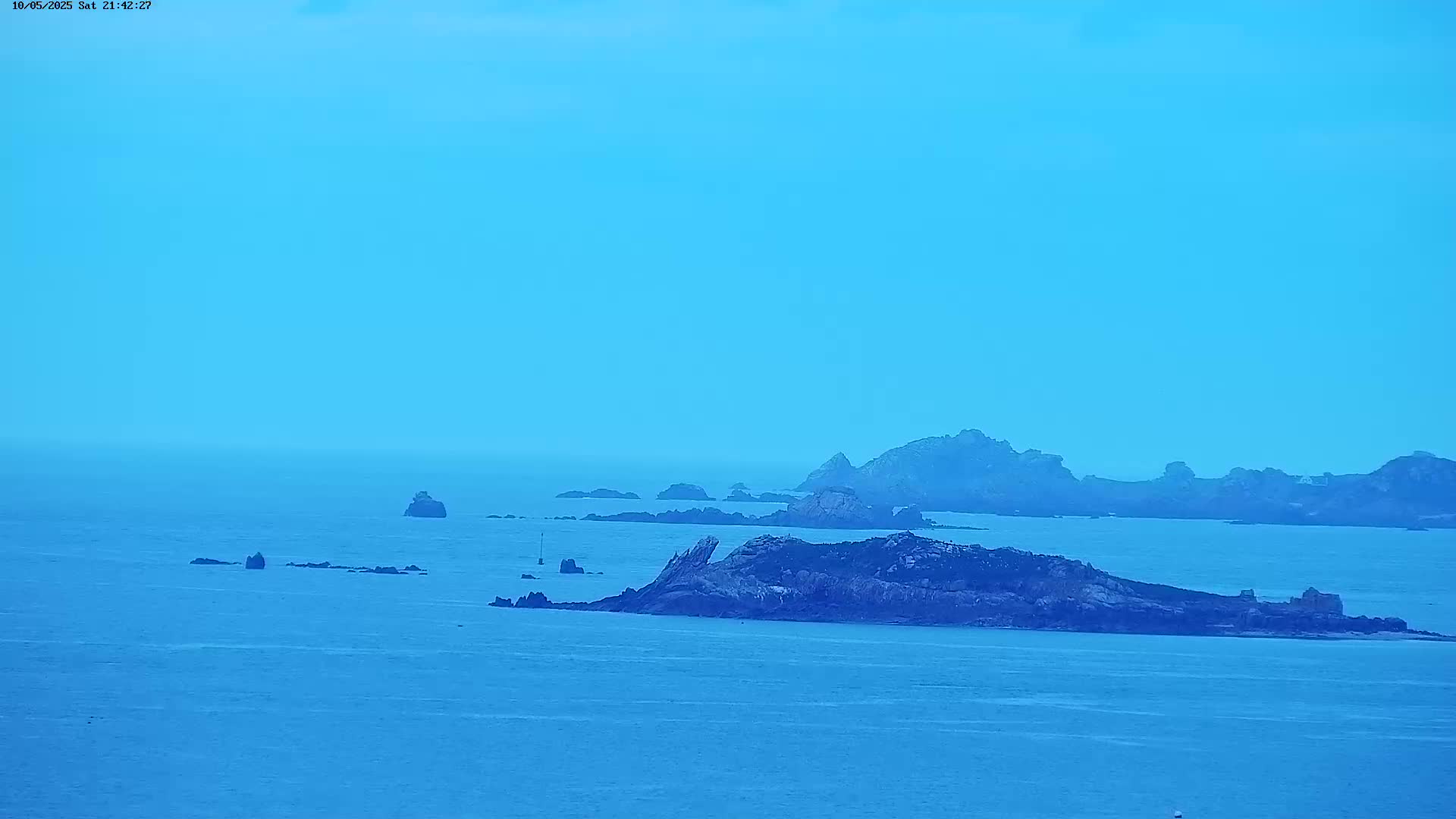A hazy, light-blue sky overlooks a calm ocean dotted with dark, rocky islands.