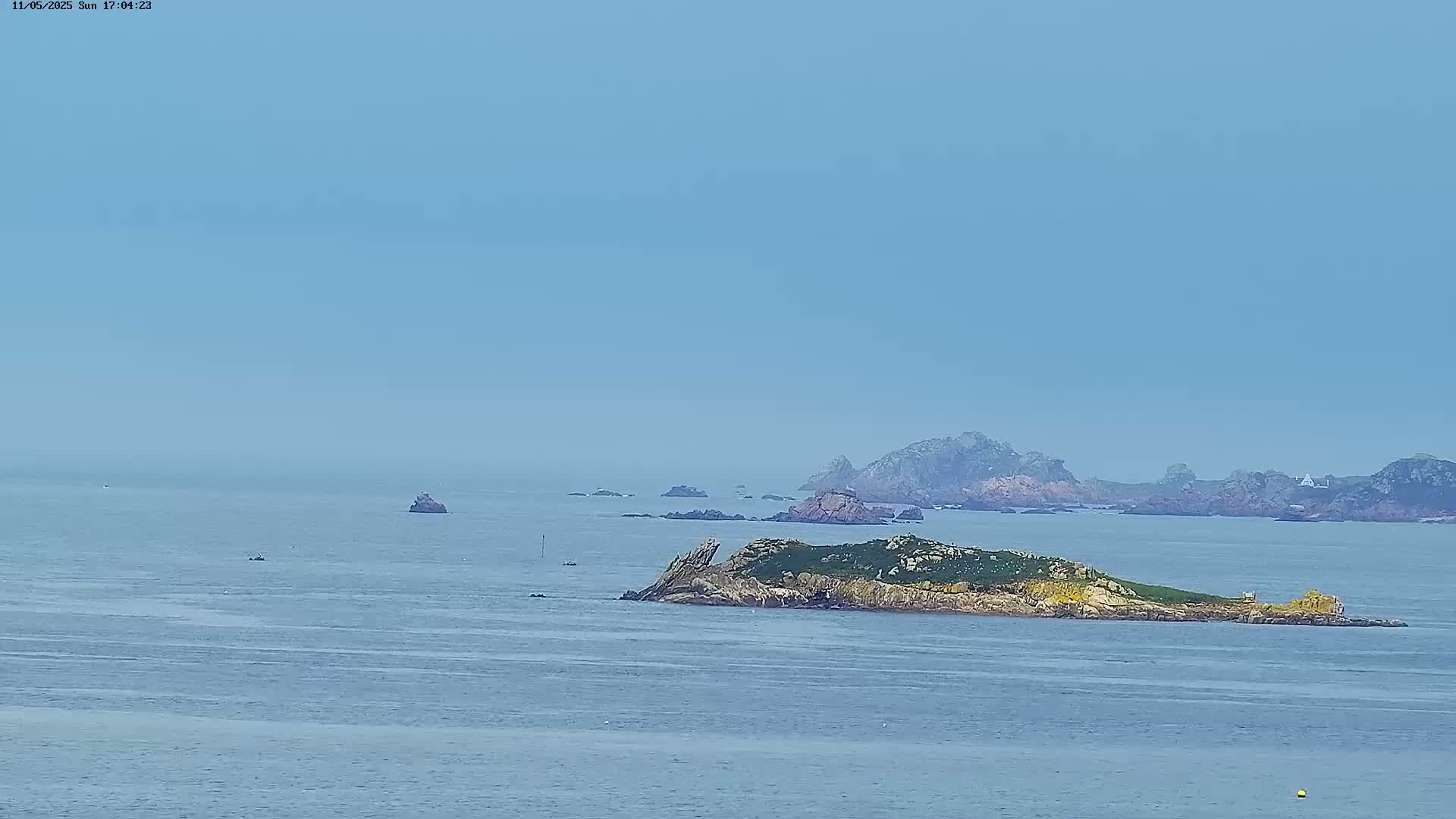 A hazy, calm ocean scene shows a small, rocky island with green vegetation, other rocky outcrops in the distance, and a few boats.