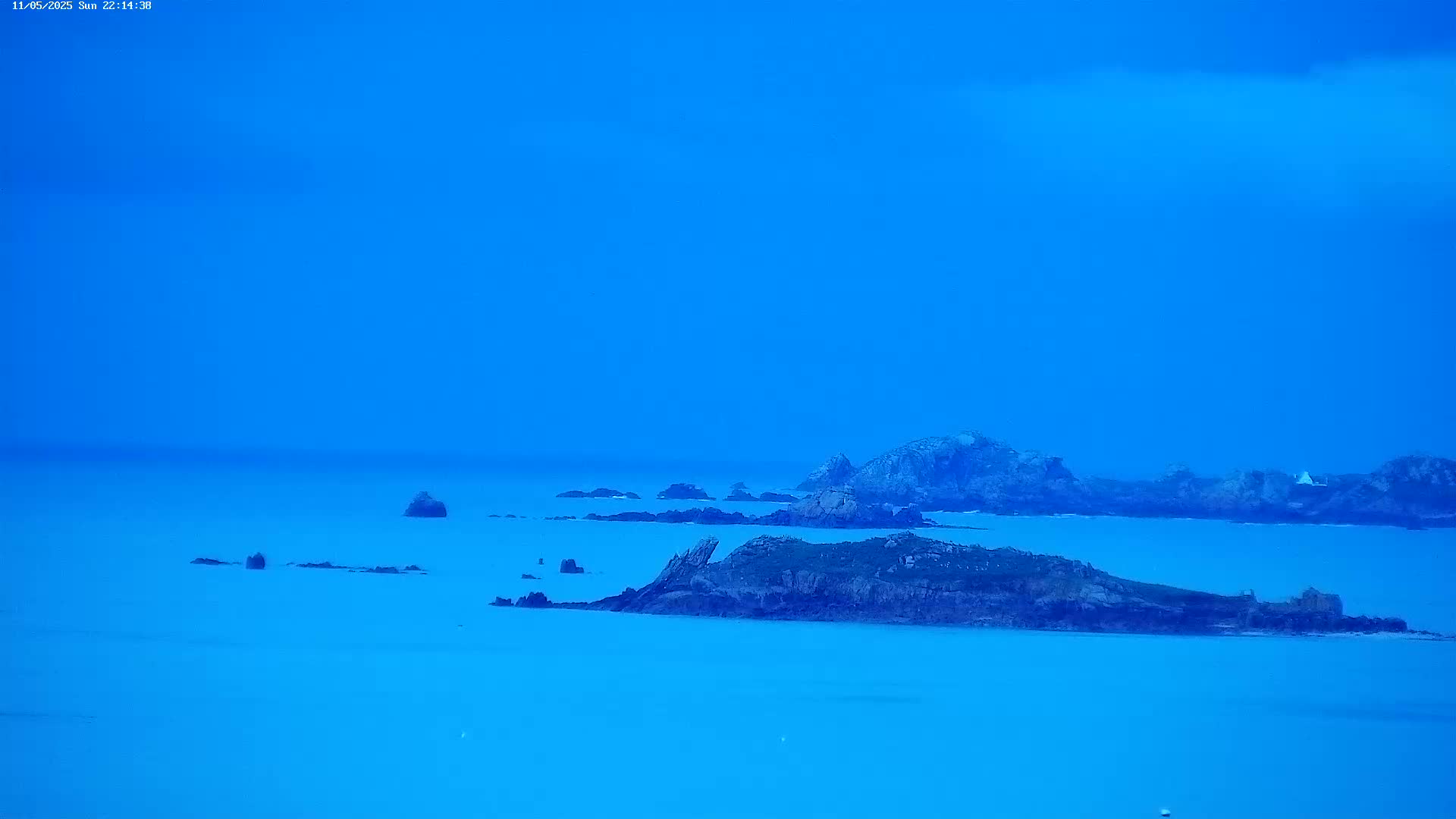 Dark, rocky islands are scattered across a calm, bright blue sea under a clear, blue sky.
