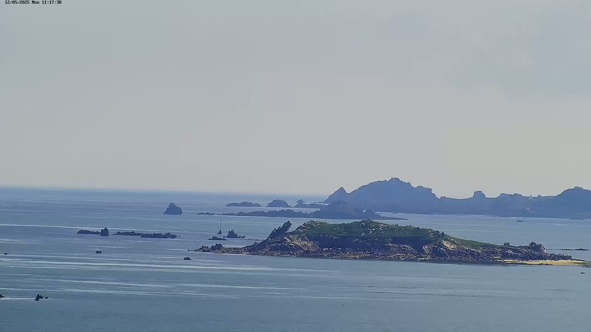 A hazy, bright day reveals a rocky island with green vegetation, surrounded by smaller rocky outcrops and a calm sea stretching to a distant shoreline.