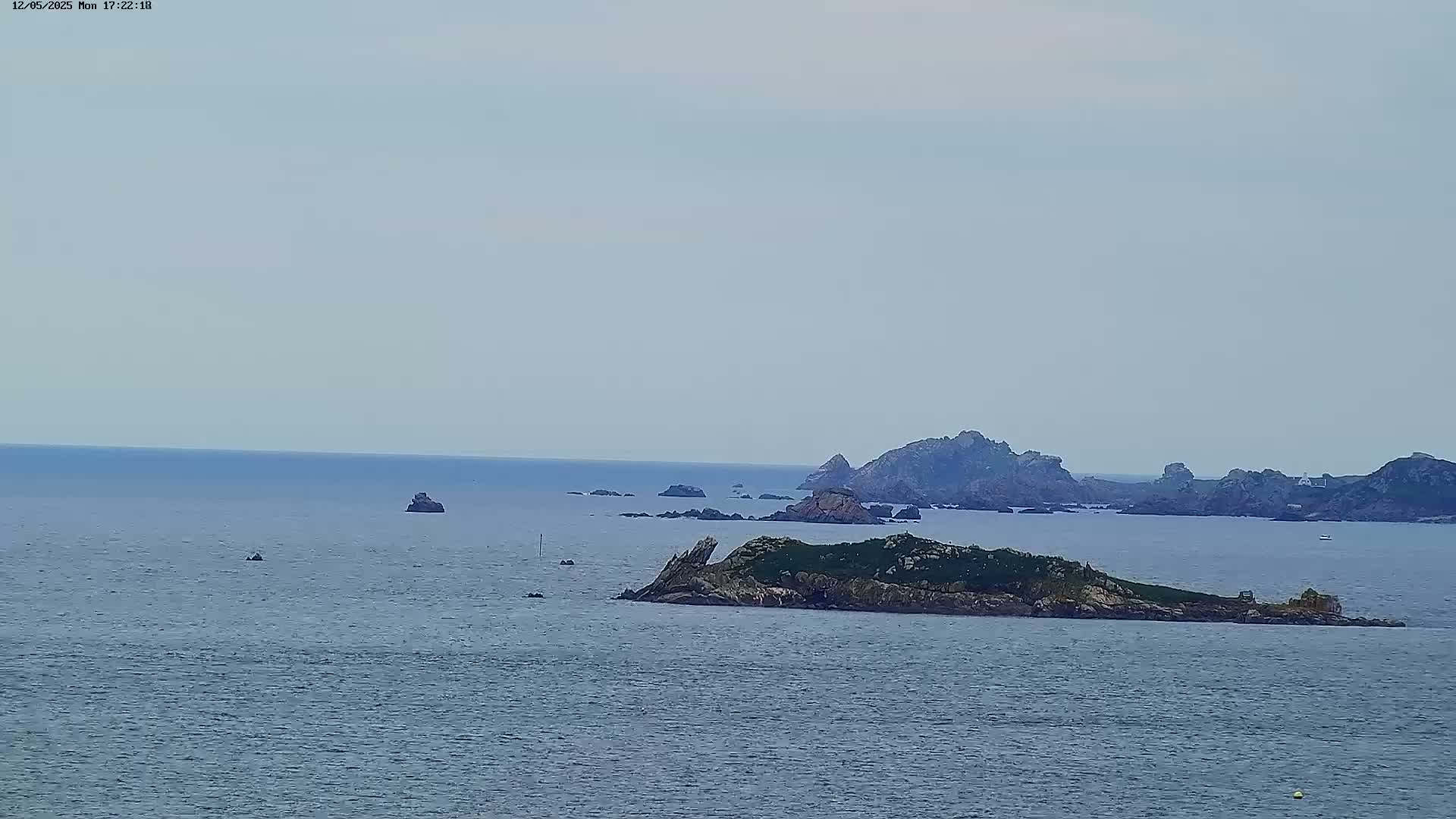 A calm, overcast day reveals a rocky island in the foreground, with additional rocky outcrops and a distant coastline visible across a tranquil sea.