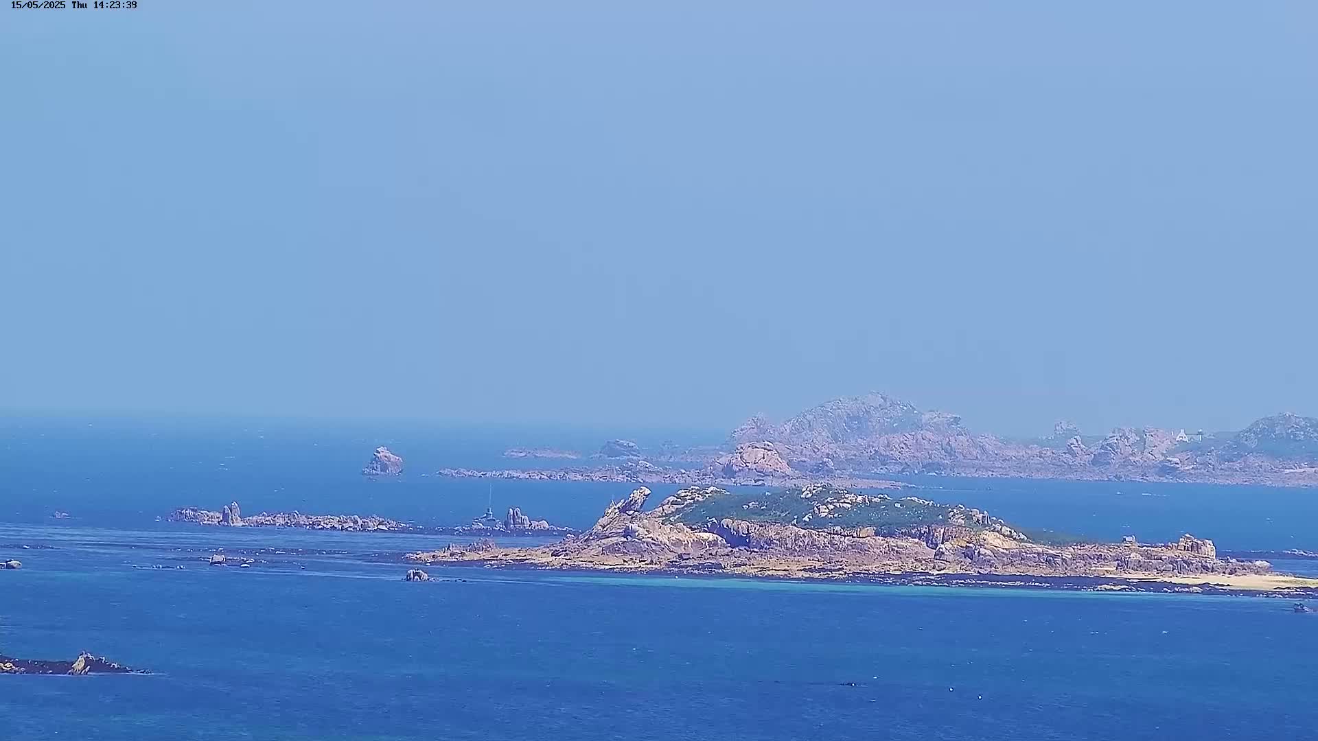 A rocky island with sparse vegetation sits in a calm, blue ocean under a hazy sky.