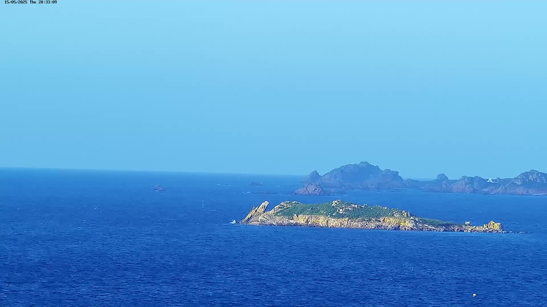 A small, vegetated island sits in a calm, deep blue ocean under a clear, light blue sky, with larger rocky islands visible in the distance.