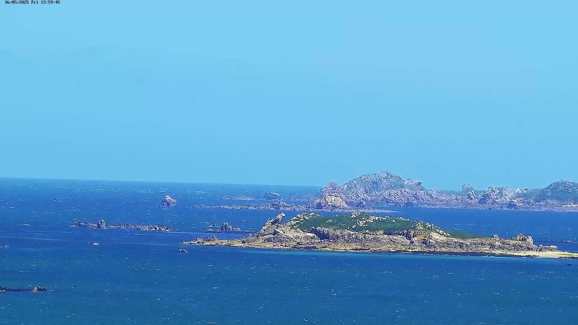 A rocky island with sparse vegetation sits in a blue sea under a clear blue sky with a light wind.