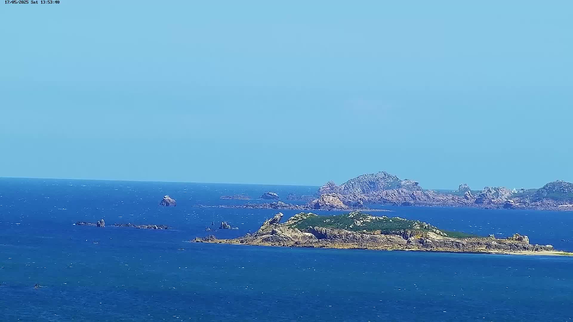 A rocky island with green vegetation sits in a calm, blue ocean under a clear, light blue sky.