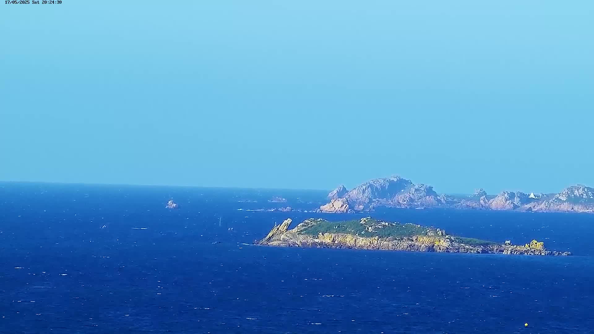 A distant view of several rocky islands in a calm, blue ocean under a clear, light blue sky.