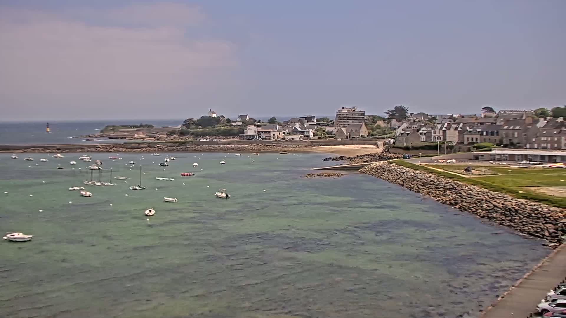 A calm, sunny day reveals a harbor filled with numerous small boats, a stone seawall, and a small coastal town nestled against a rocky shoreline.