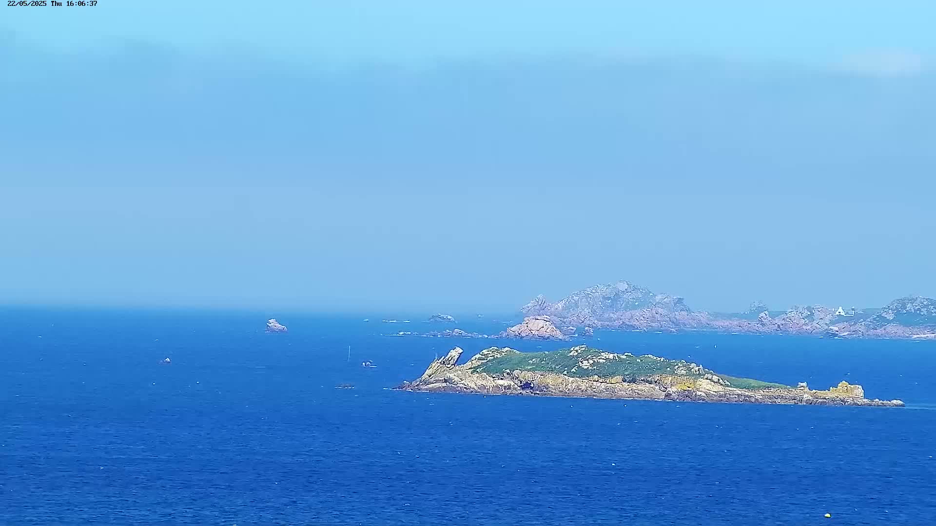 A rocky island with green vegetation is visible in the middle of a calm, blue ocean under a clear, light-blue sky, with other smaller islands and landmasses in the distance.