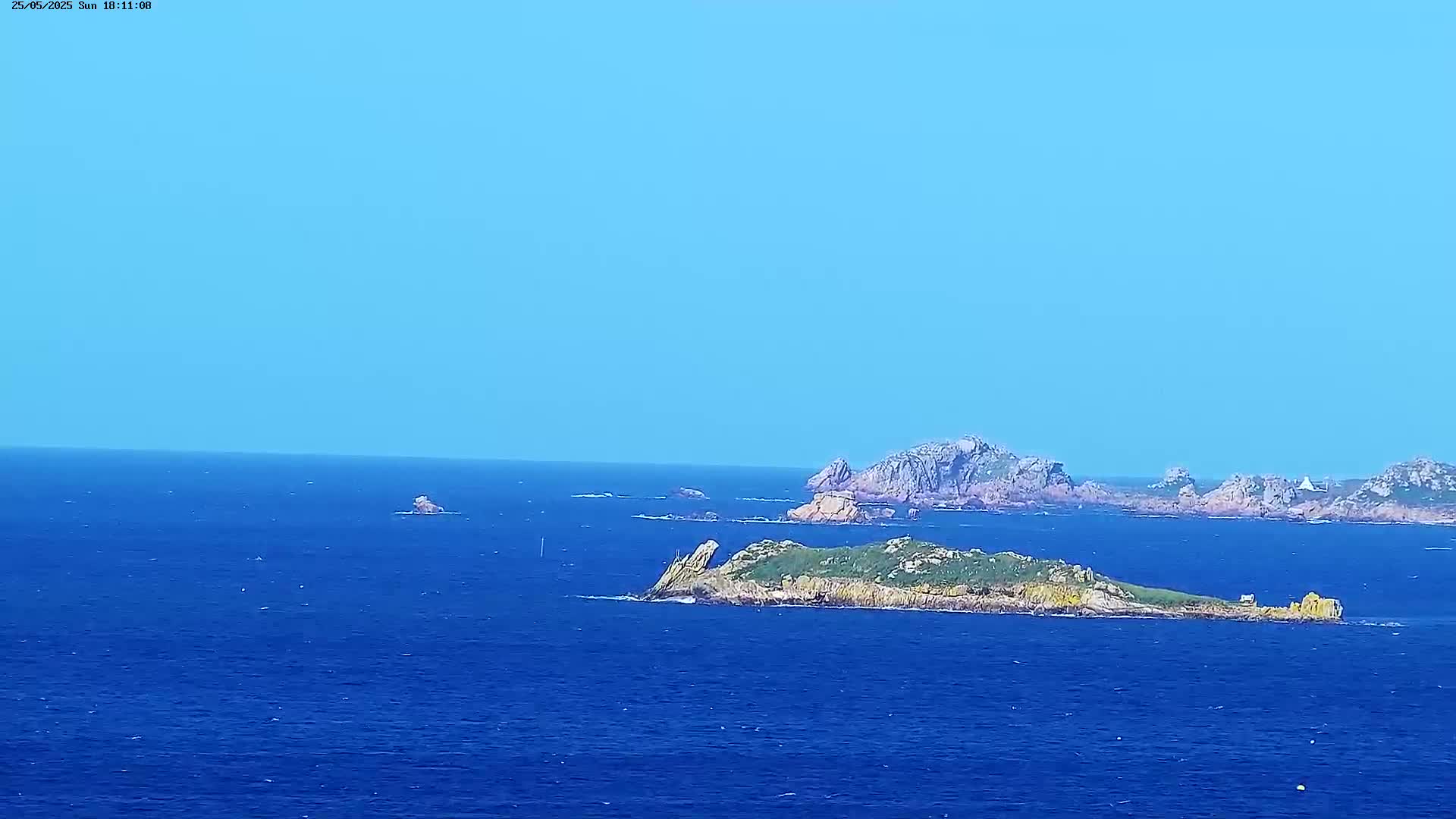 A small, green-topped island sits in a vast, deep blue ocean under a clear, light blue sky.