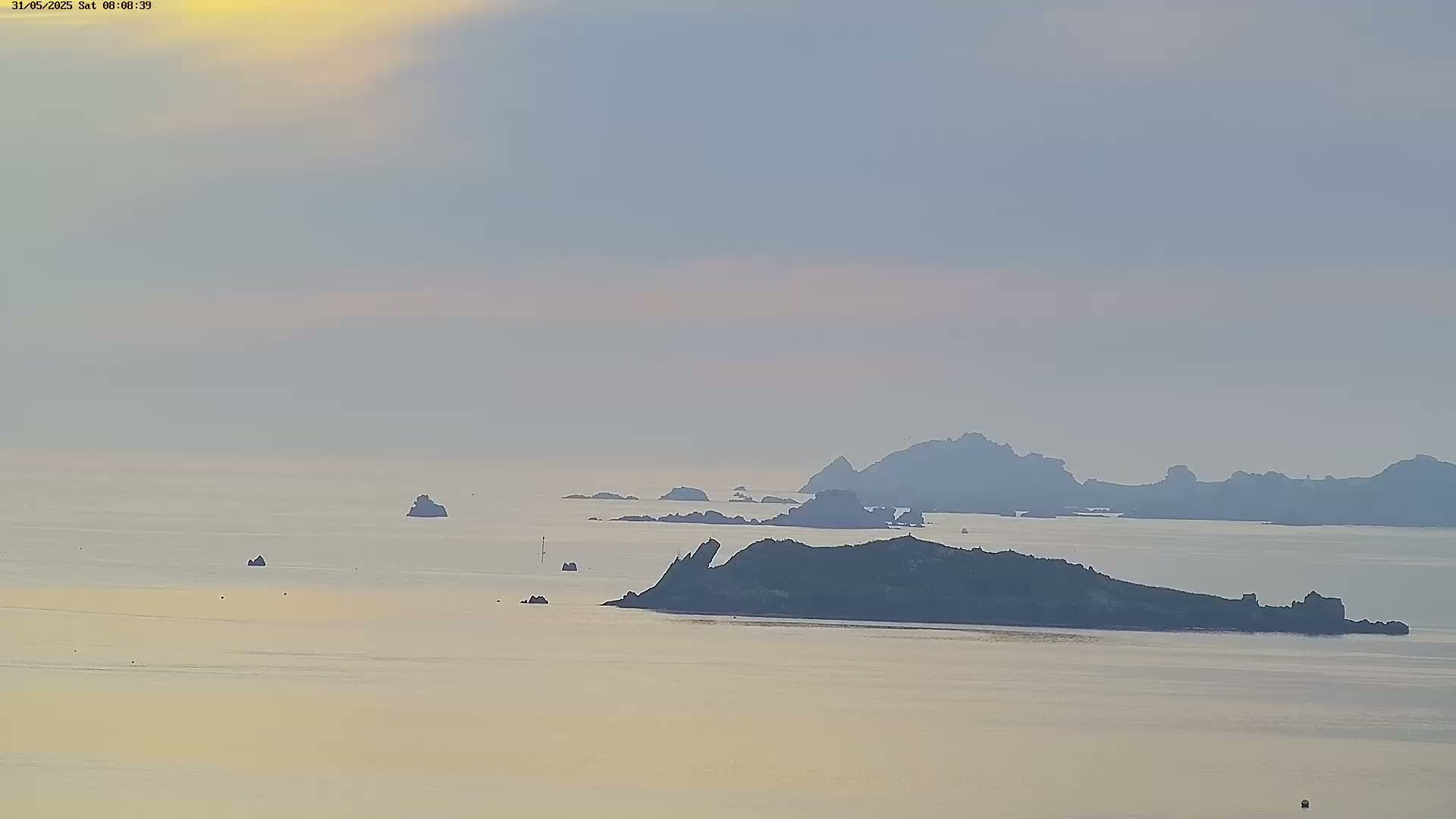 A hazy, calm sea with several small islands and rock formations under a pale, sunrise sky.