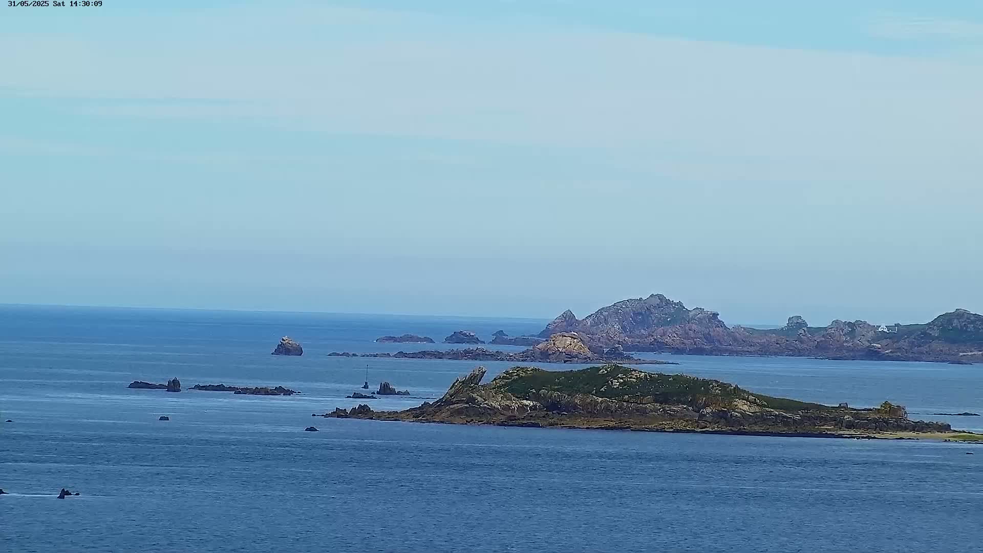 A distant view of a rocky island chain and several smaller islets in a calm ocean under a clear, light-blue sky.