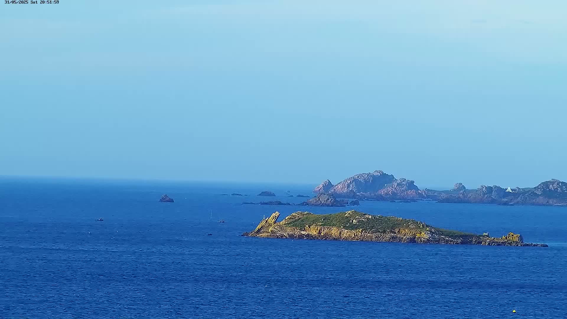 A picturesque, rocky island sits in a calm, blue ocean under a clear, light blue sky, with other small islands and rocky outcrops visible in the distance.
