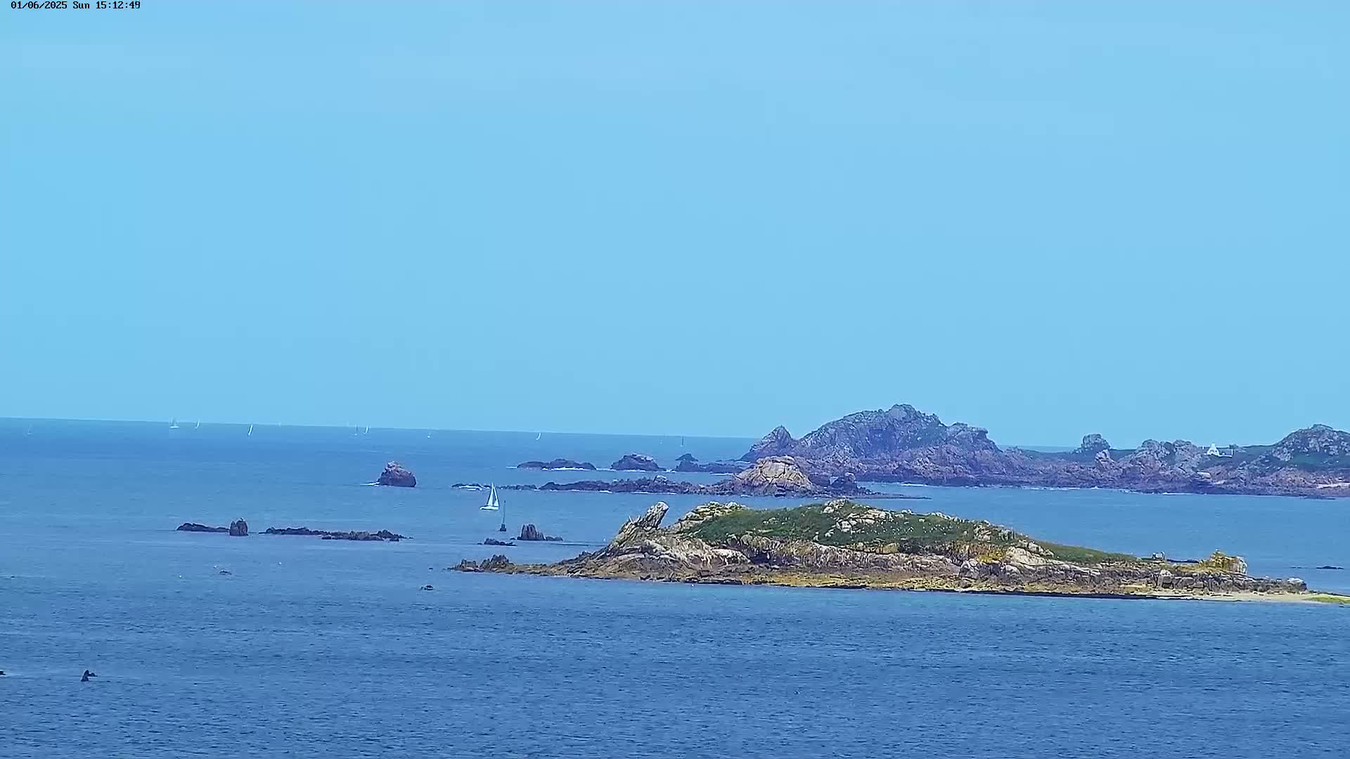 A small, grassy island with rocky shores sits in a calm, blue ocean under a clear sky, with several other rocky outcrops and a few distant sailboats visible.