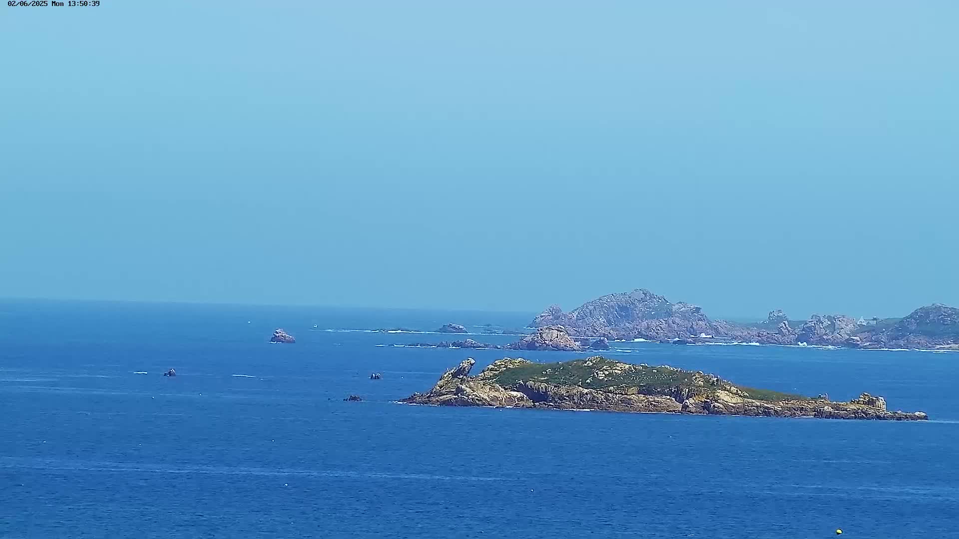 A small, vegetated island sits in a calm, blue ocean under a clear, bright sky, with other rocky outcrops visible in the distance.