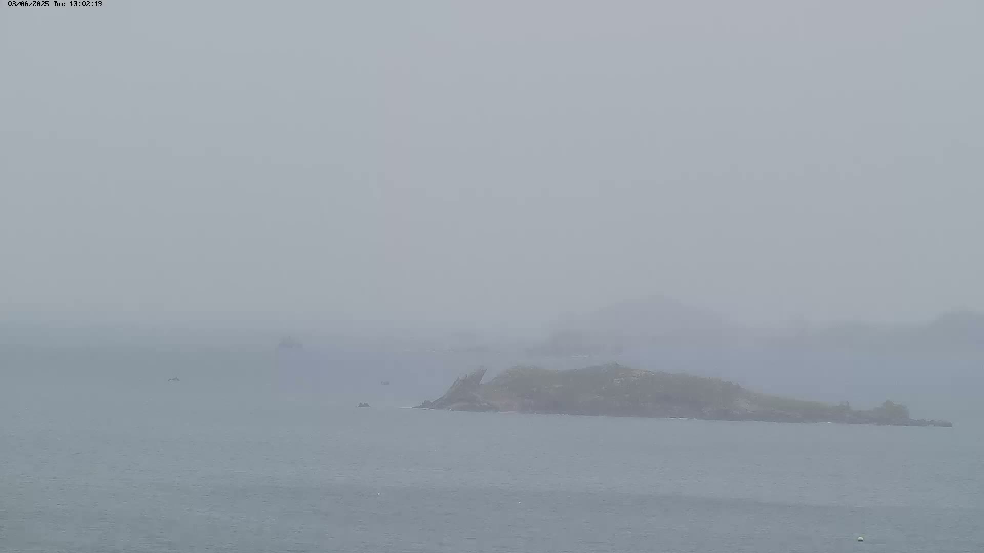A hazy, overcast day shows a small, dark island in the middle of a calm sea, with a few small boats visible in the distance.