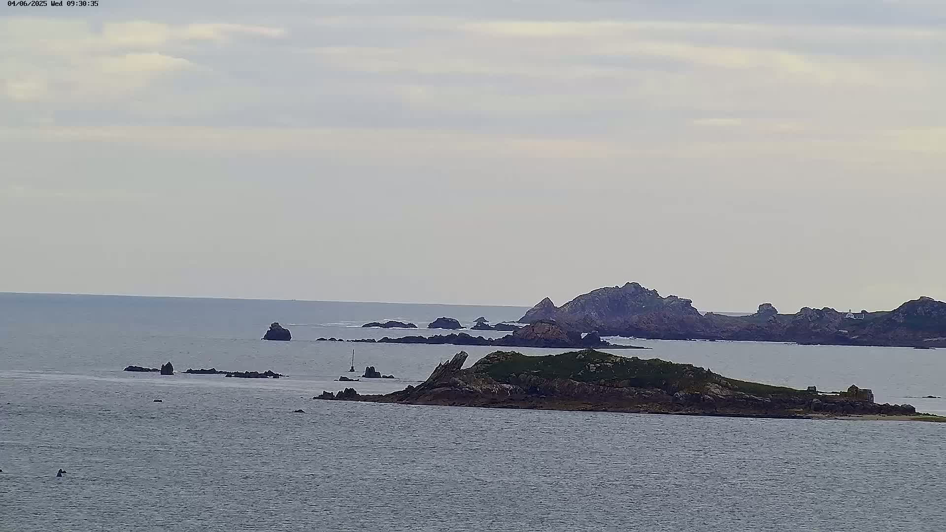 A calm, overcast day reveals a rocky coastline with several small islands scattered across a gray sea.