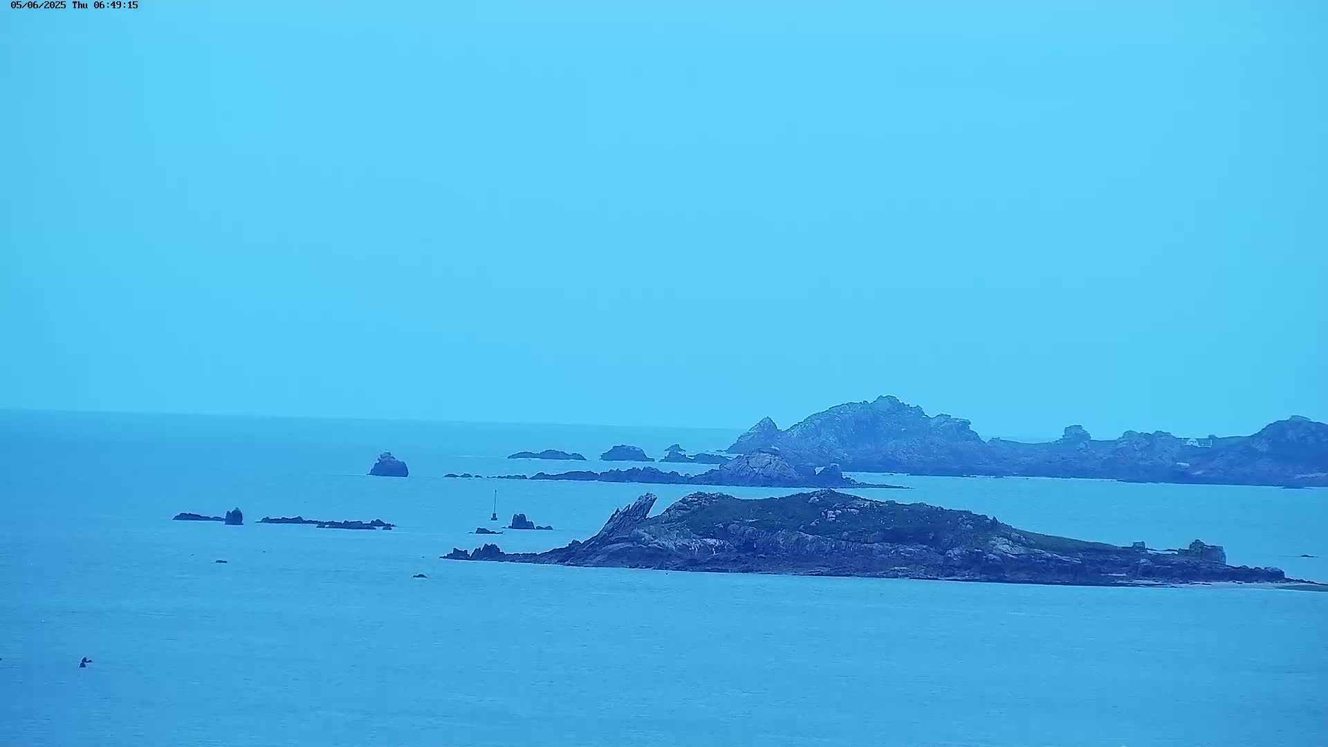 A tranquil ocean scene under a pale blue sky shows several rocky islands and outcroppings scattered across the water.