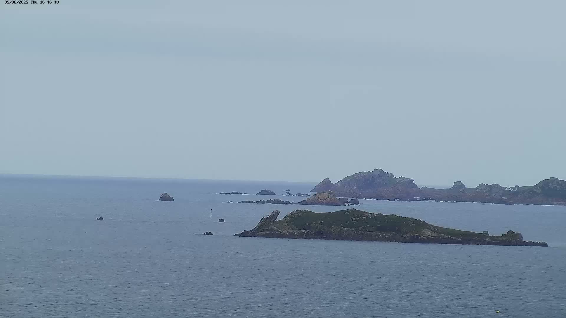 A wide shot of a rocky, low-lying island surrounded by a calm, grey sea under an overcast sky.