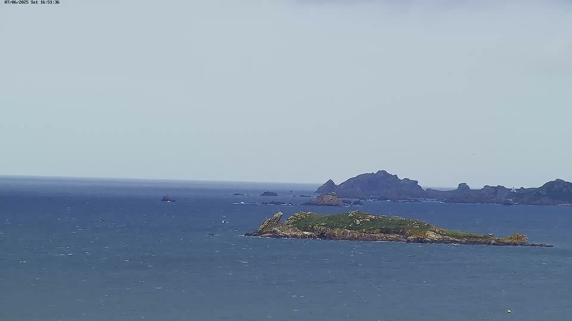 A small, grassy island sits in a calm, gray sea, with larger rocky islands visible in the distance under a cloudy sky.