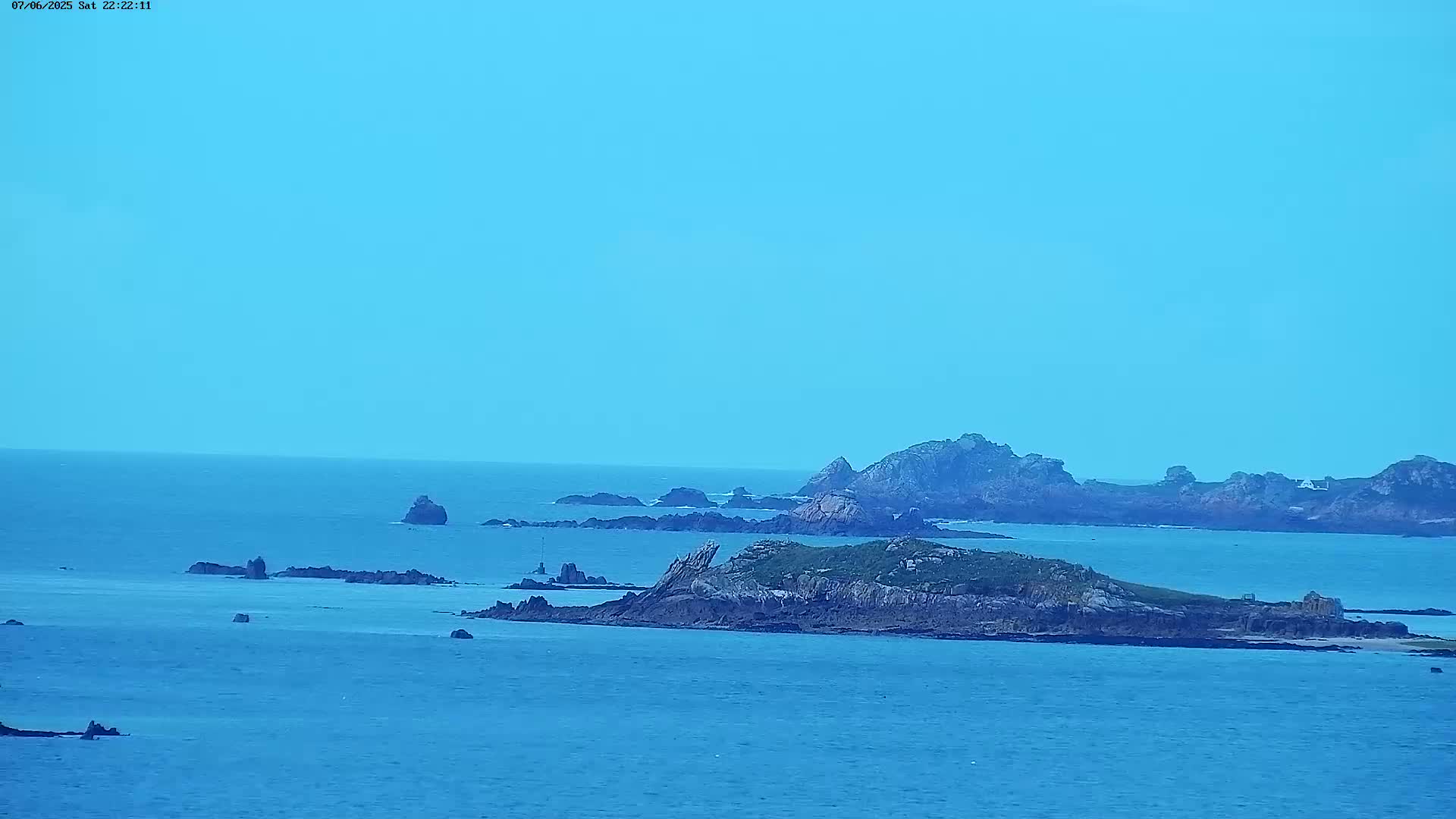 A tranquil ocean scene under a clear, light-blue sky features several rocky islands and islets scattered across a calm expanse of water.