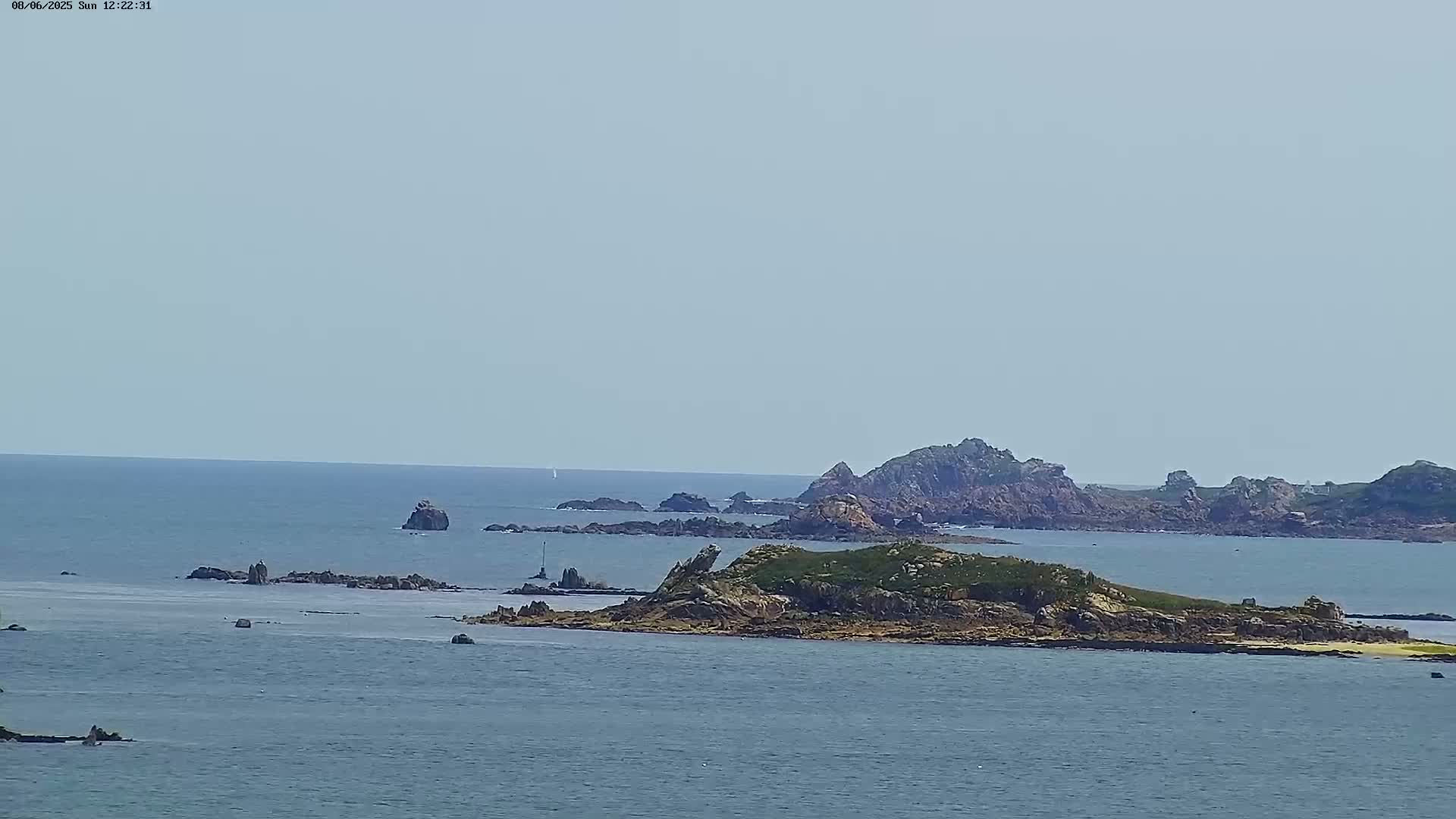 A small, rocky island with sparse green vegetation is visible in a calm, blue ocean under a clear sky, with other rocky outcrops and landmasses in the distance.