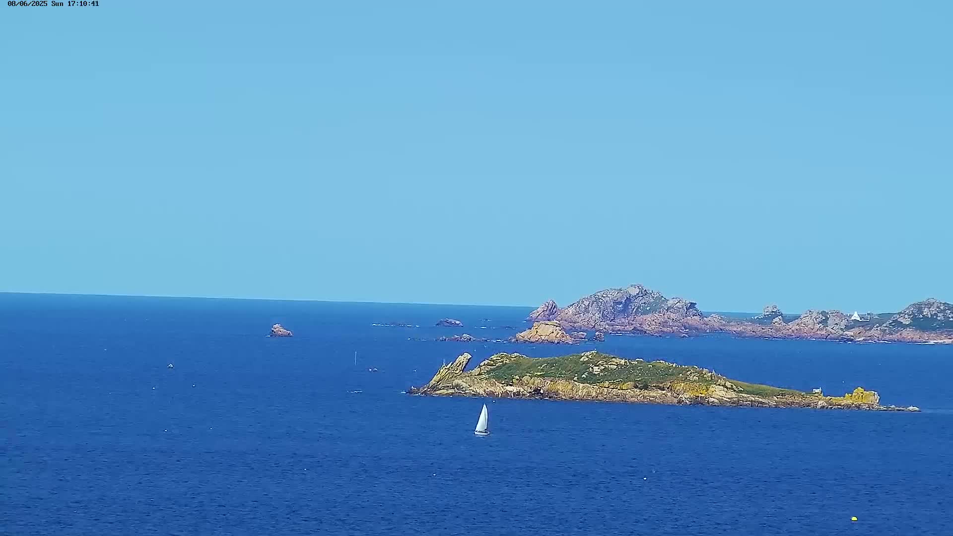 A sailboat is visible on a calm, blue ocean near rocky islands under a clear, blue sky.