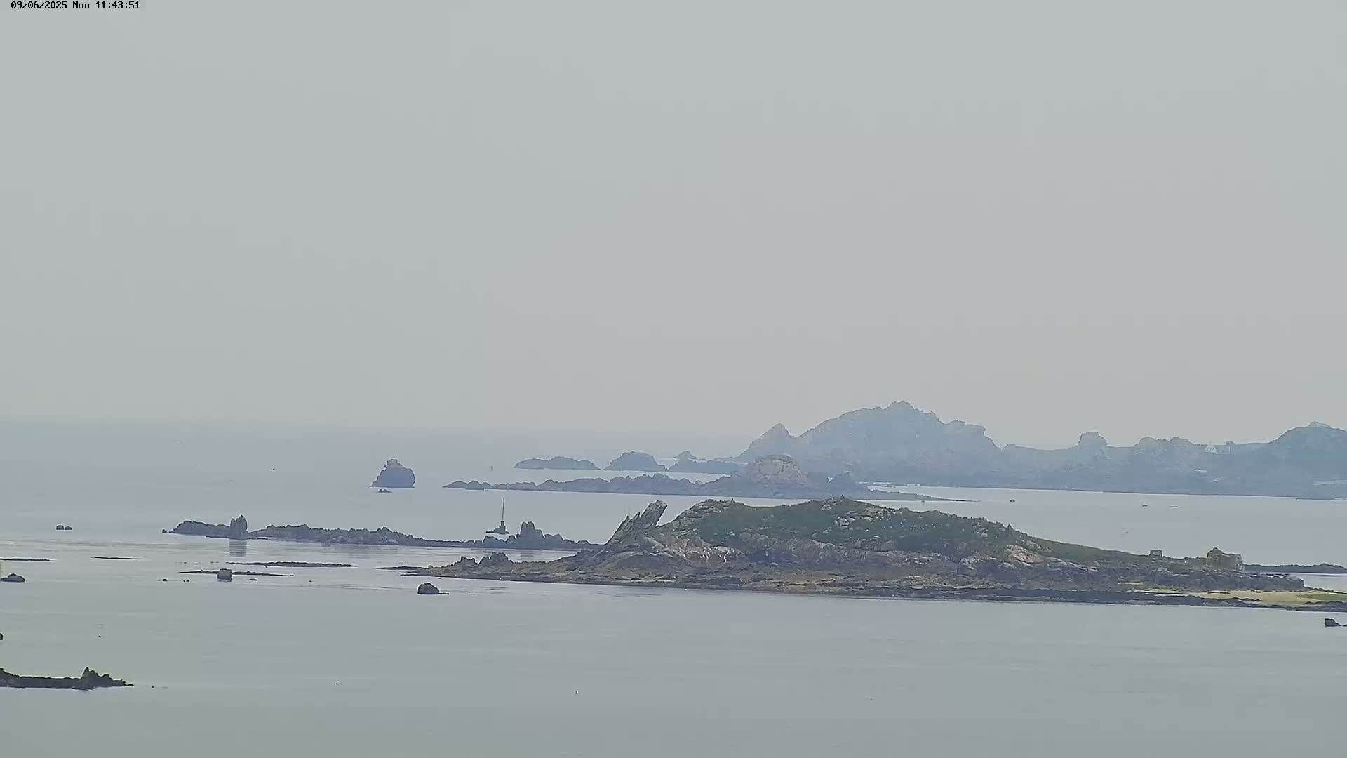 A hazy, overcast day reveals a rocky island and coastline, with several smaller rock formations scattered in the surrounding calm water.