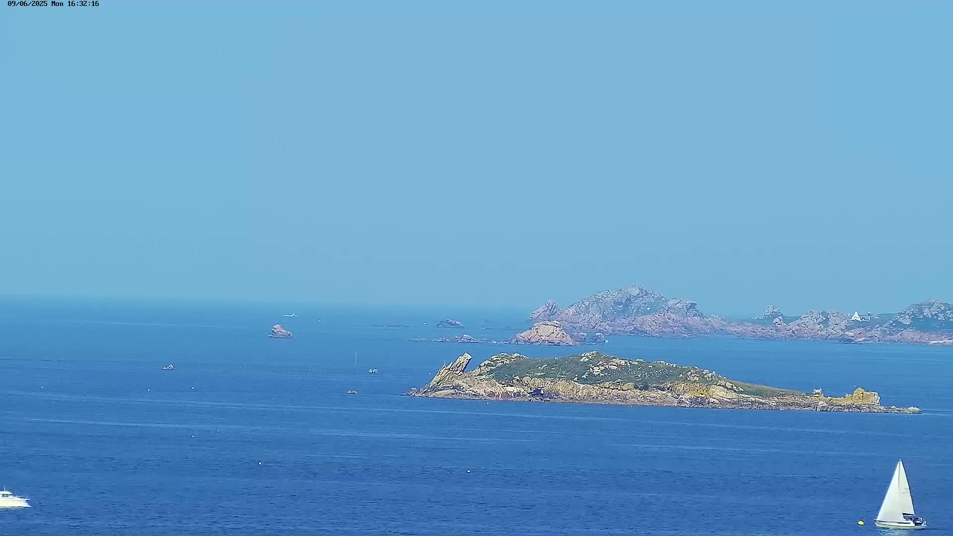 A sailboat and several small boats are visible on a calm, blue ocean with rocky islands in the distance under a clear, light blue sky.
