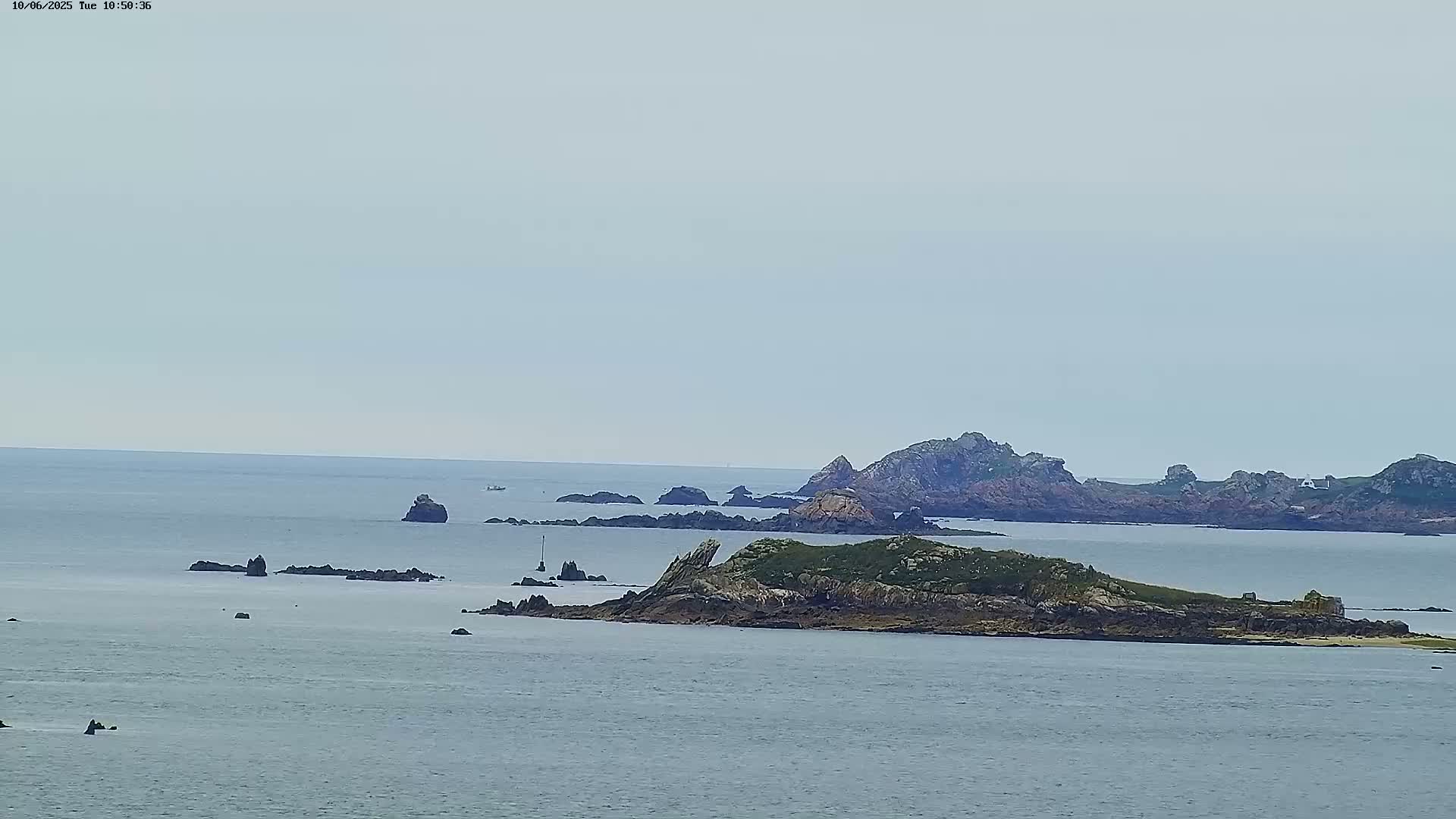 A calm, gray day reveals a rocky, low-lying island surrounded by numerous smaller rocks and islets in a vast, tranquil sea.