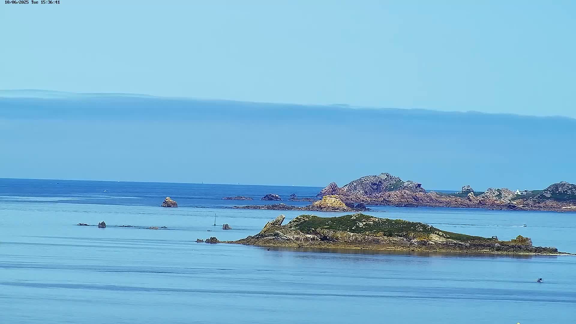 A tranquil ocean scene under a clear, light-blue sky shows several rocky islands and islets, some with sparse vegetation.