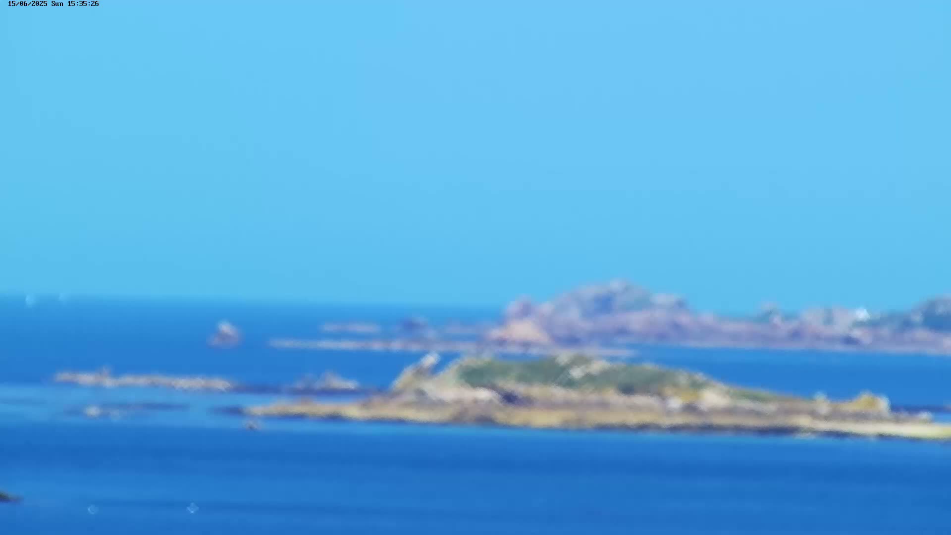 A blurry, long-distance view of rocky islands and a calm, blue ocean under a clear blue sky.