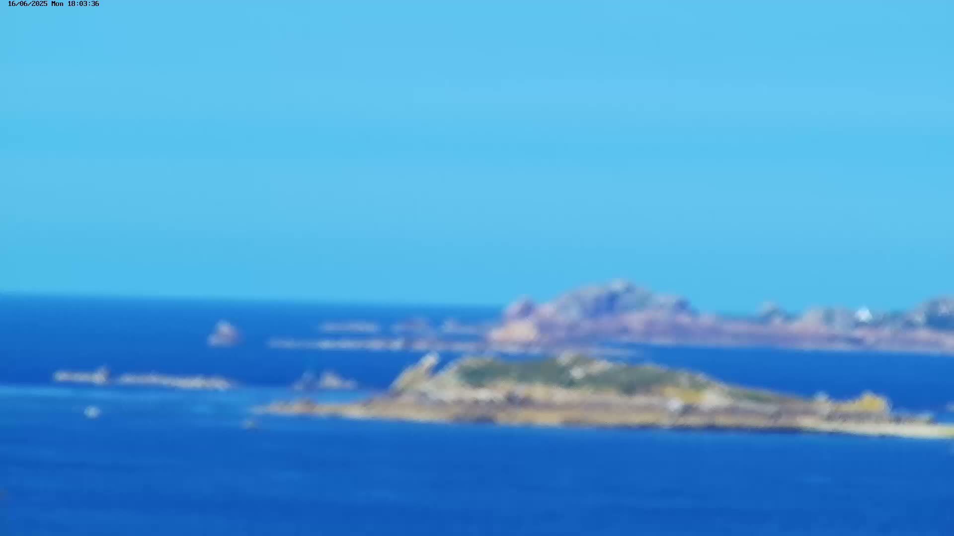 A blurry, long-distance view shows a rocky island in a calm, blue ocean under a clear, bright blue sky.