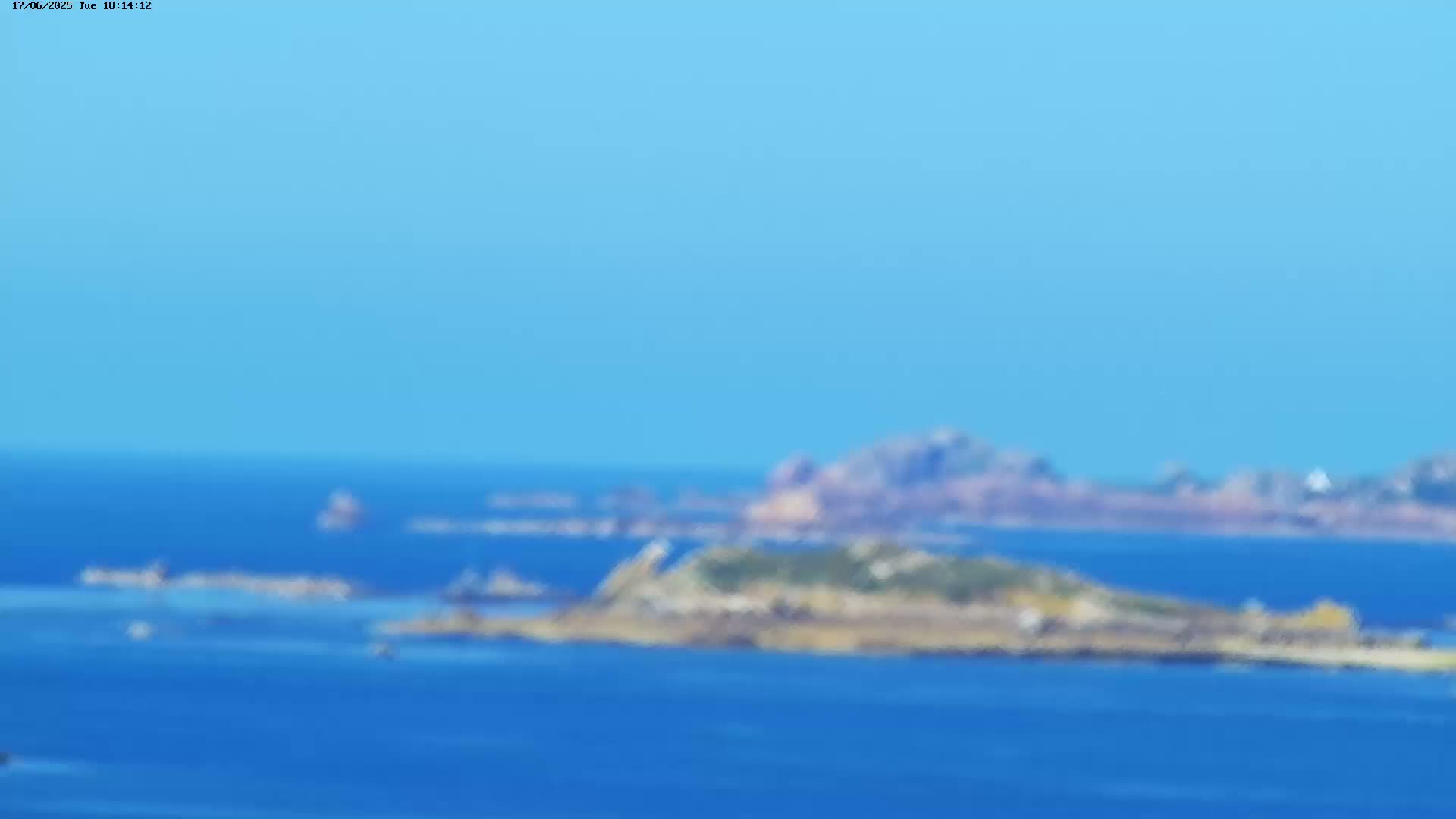 A blurry long shot shows a rocky island in a calm blue ocean under a clear blue sky.