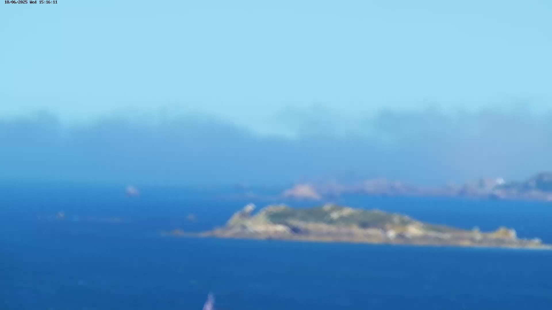 A hazy, sunny day shows a view of a small, low-lying island in a calm blue ocean with other landmasses visible in the distance.