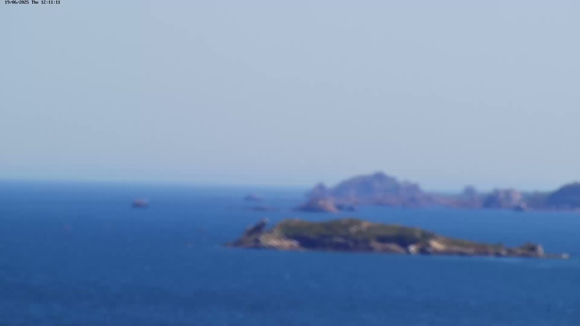 A blurry, long-distance view of several small, dark islands in a calm, blue ocean under a clear sky.