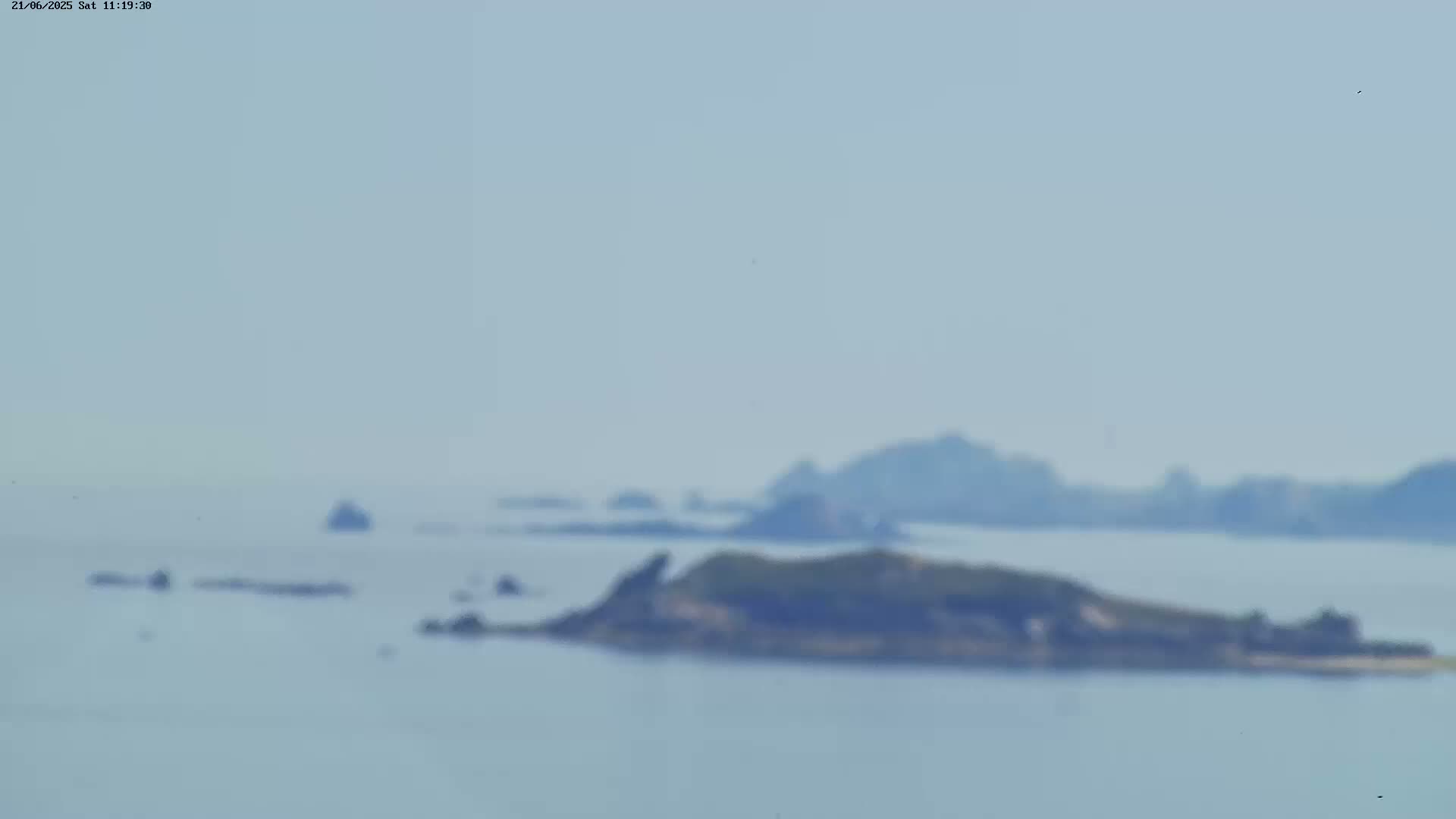 A hazy, light-blue ocean scene shows several small, dark islands on a calm sea under a mostly clear sky.