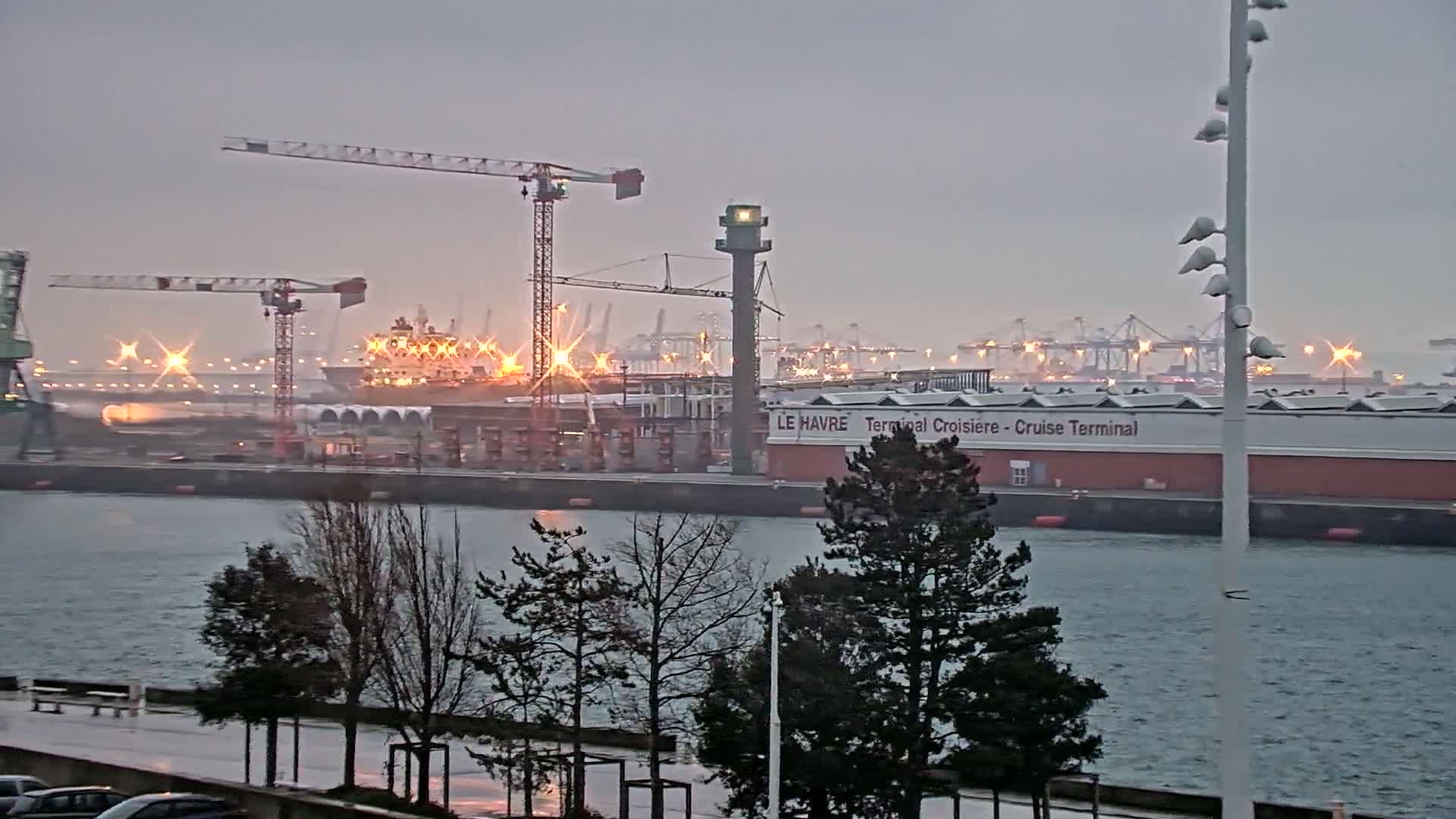A ship is docked at a harbor under an overcast sky, with several cranes and industrial buildings visible in the background.