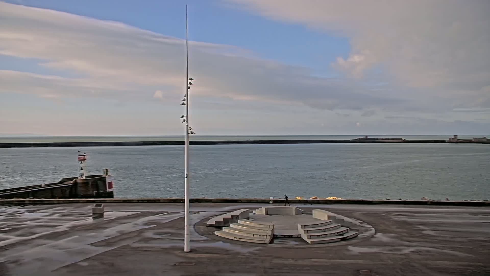 A tall flagpole stands in a wet, paved area with a circular amphitheater-like structure near a calm body of water under a partly cloudy sky.