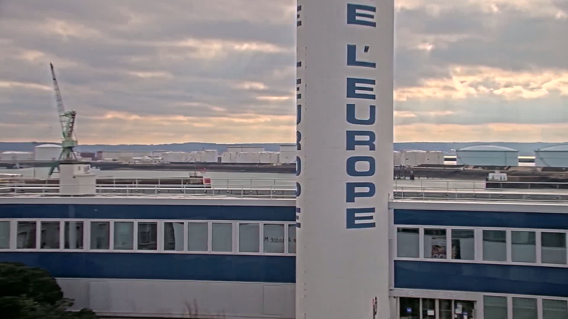 A large white tower with "L'EUROPE" written vertically on it stands beside a blue building overlooking a body of water with industrial storage tanks and a crane in the distance under a cloudy sky.