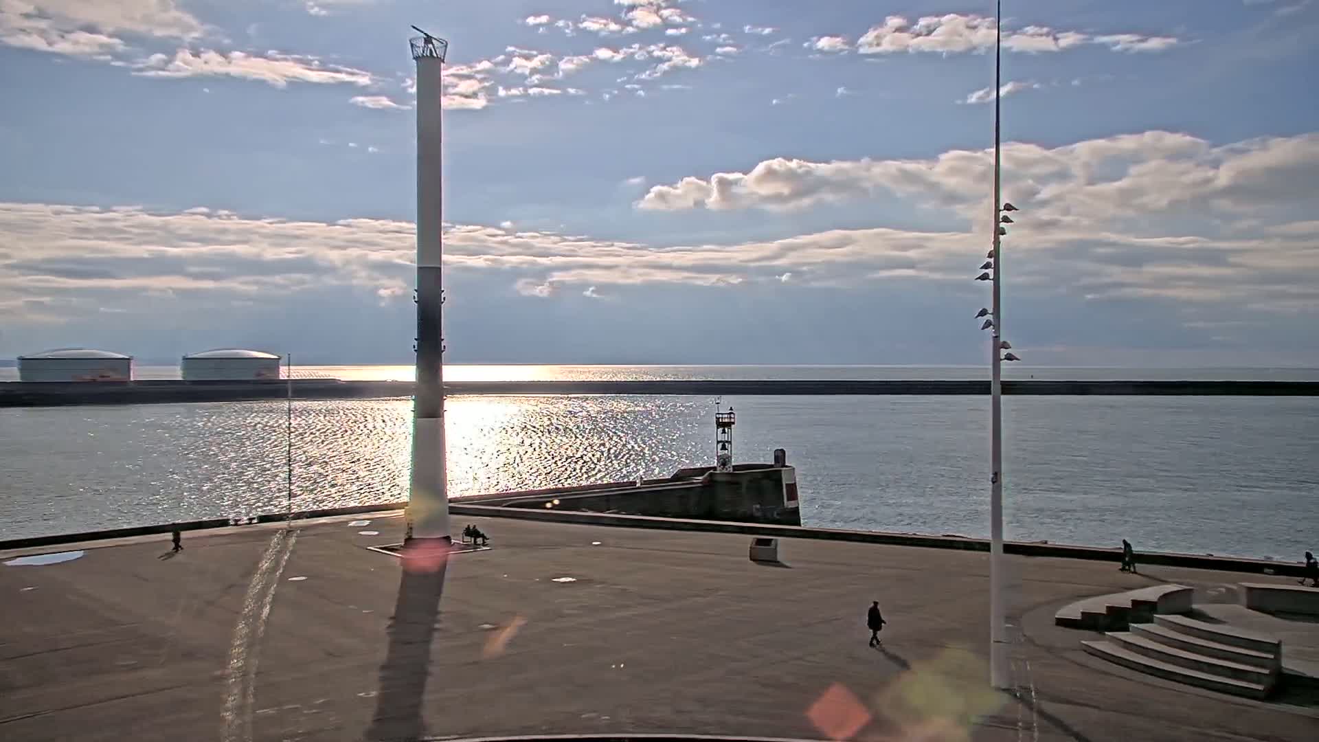 A mostly sunny day with some clouds shows a harbor area with a tall, slender tower, a smaller lighthouse, and a few people walking on a paved area next to calm water.