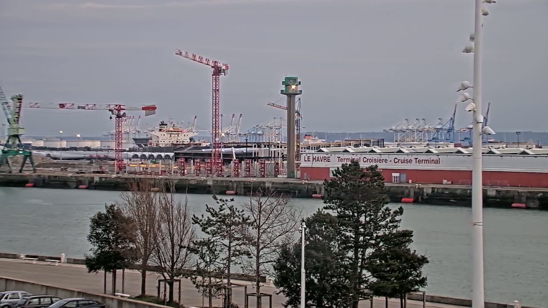 A harbor scene under an overcast sky shows construction activity near a cruise terminal, with several cranes and a ship visible.