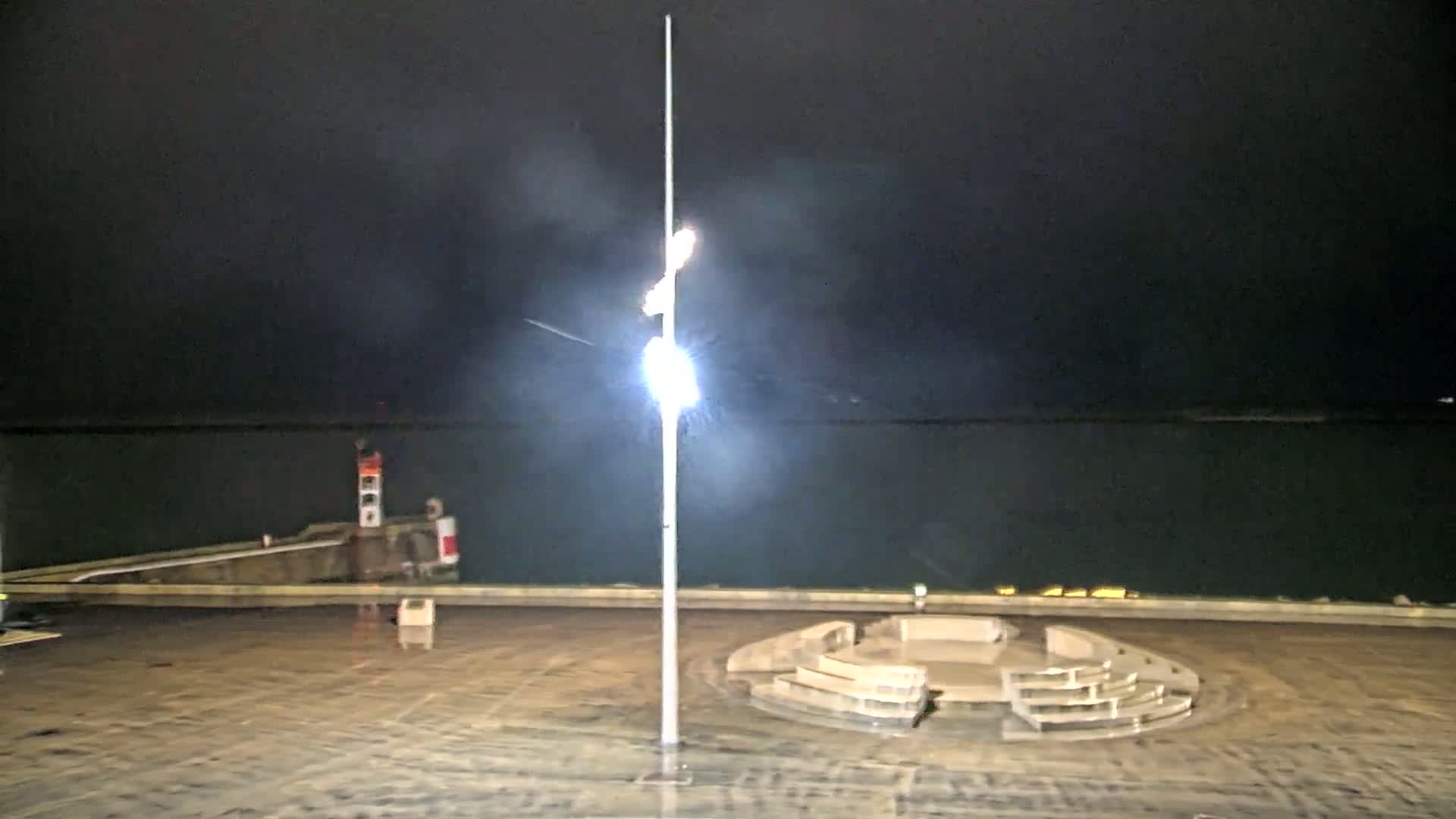 A tall light post stands in a wet, nighttime waterfront plaza near a small lighthouse and a dark body of water.