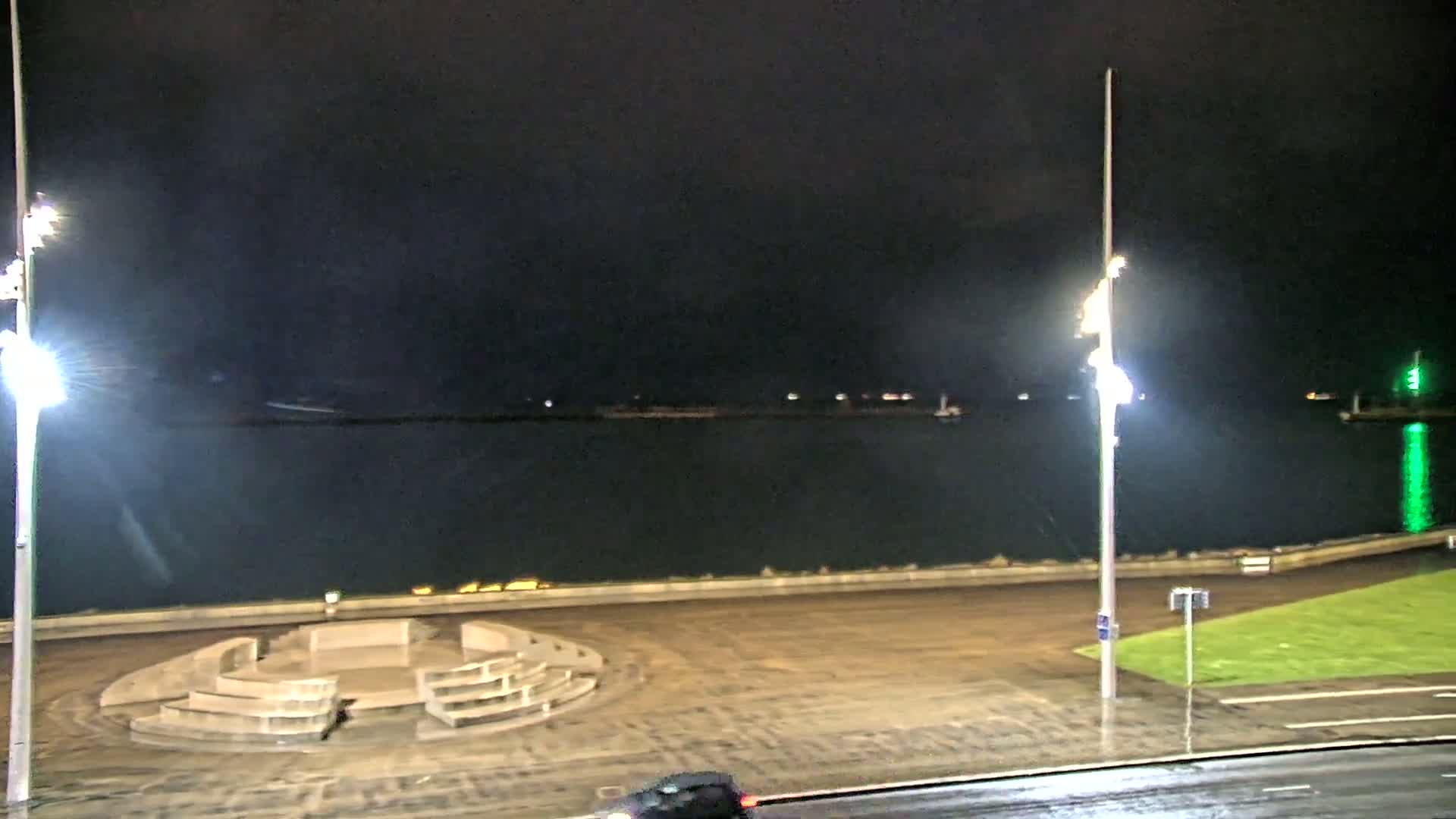 A nighttime view of a waterfront area shows a dark body of water, a concrete seating area, and a wet road under bright lights.