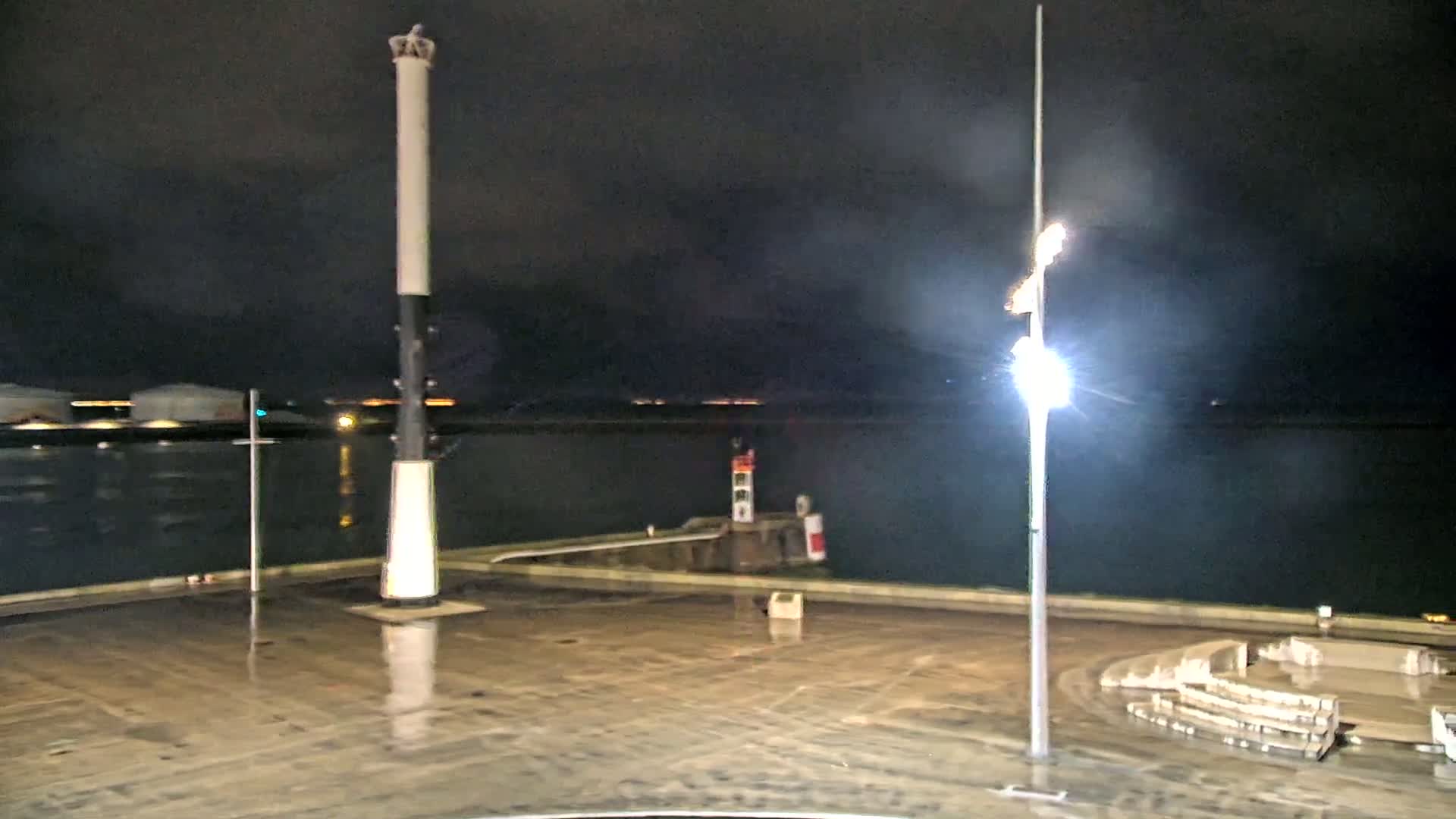 A wet, nighttime harbor scene shows a tall, slender lighthouse, a smaller harbor light, and a tall lamppost, all on a concrete area beside dark water.