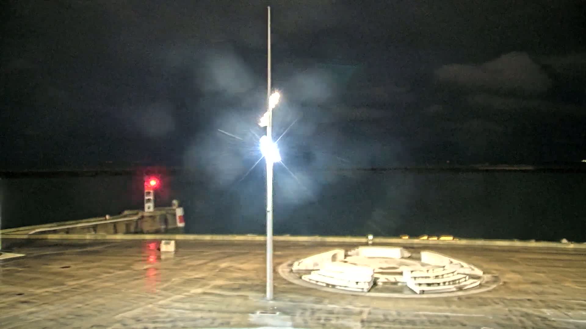 A brightly lit lamppost stands in a mostly empty plaza at night near a body of water, with a red light visible in the distance under a hazy, dark sky.